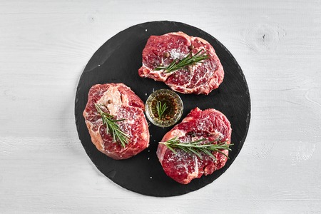 Fresh raw beef steaks with herbs, garlic, olive oil, pepper, salt and rosemary on black board: Tenderloin, Striploin, Rib Eye on white background. Top view. Copy space. Still life. Flat layの写真素材