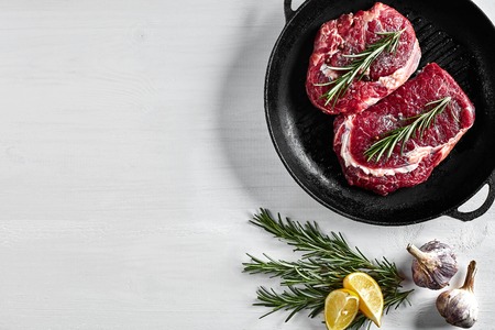 Raw Steak in grill frying pan with herbs and oil on white background, top view, place for textの写真素材