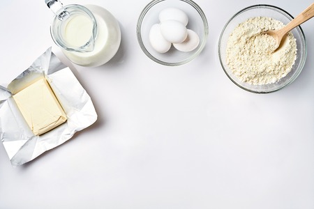 Wheat flour in the glass bowl, cream in a glass jar, butter, chicken eggs - ingredients for the dough, on a white background. Top view. Copy space. Still life. Flat layの写真素材