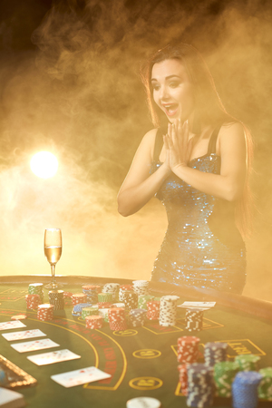 Young beautiful woman poses near poker table in luxury casino. Passion, cards, chips, alcohol, dice, gambling, casino - it is as female entertainment. Smoke background.の写真素材