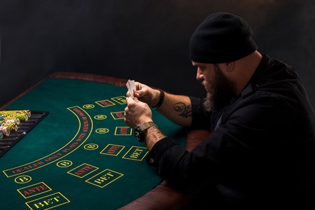 Serious bearded man sitting at poker table and holding cards isolated on black. Copy spaceの写真素材