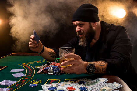 Bearded man with cigar and glass sitting at poker table in a casino. Gambling, playing cards and roulette.の写真素材
