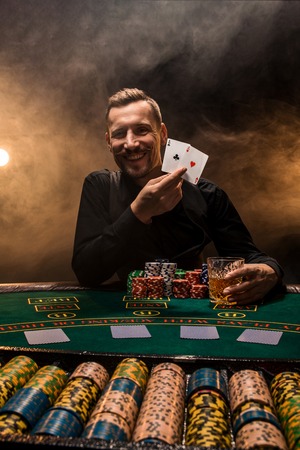 Handsome poker player with two aces in his hands and chips sitting at poker table in a dark room full of cigarette smoke.の写真素材