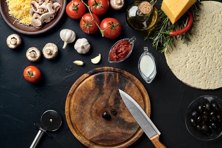 Cutting wooden board with traditional pizza preparation ingredients: cheese, tomatoes sauce, olives, olive oil, pepper, spices. Black texture table background. Top view. Still life. Copy space. Flat layの写真素材