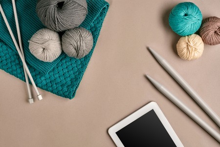 Grey and turquoise knitting wool, knitting needles and a tablet with a black screen on beige background. Knitting as a kind of needlework. Colorful balls of yarn and knitting needles. Top view. Still life. Copy space. Flat layの写真素材