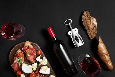 Cheese appetizer selection or wine snack set. Red wine, cheese, cherry tomato, bread and prosciutto on wooden board over black backdrop, top view, copy spaceの写真素材