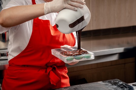 A woman confectioner with red uniform and white sterile gloves do a set of colorful chocolates from milk chocolate on a table.の写真素材