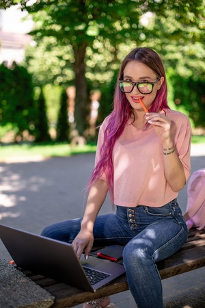 Young girl is studying in the spring park, sitting on the wooden bench and browsing on her laptop