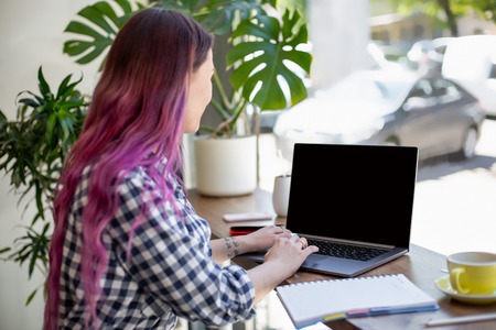 Back view of a young pink hair woman keyboarding on laptop computer with blank copy space screen while sitting in cafe.の写真素材