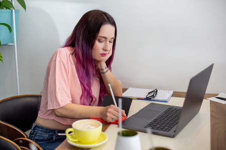 Young woman with pink hair with laptop computer sitting in cafe, intelligent female student working on net-book.の写真素材
