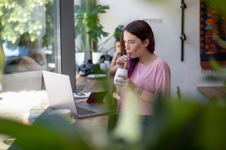 Side view of young businesswoman sitting at table in coffee shop. On table cup of coffee and laptop. In background white wall and window.の写真素材