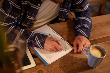 Young bearded businessman sits in cafe, home at table and writes in notebook, near lies tablet computer with black screen. Man is working, studying.の写真素材