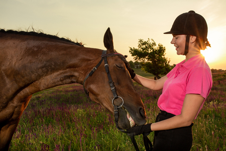 Beautiful smiling girl jockey stand next to her brown horse wearing special uniform on a sky and green field background on a sunset.の写真素材