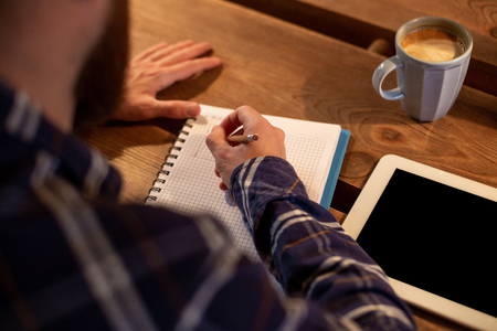 Young bearded businessman sits in cafe, home at table and writes in notebook, near lies tablet computer with black screen. Man is working, studying.の写真素材