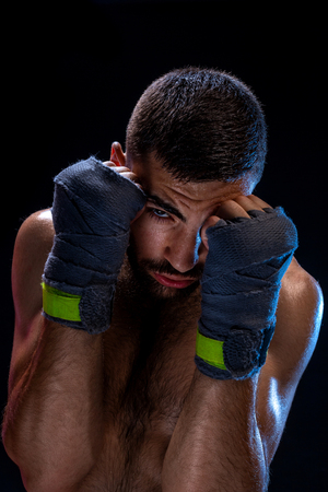 Determined young man with athletic body wearing training bandages posing in defensive position, holding pumped fists in front of his face and staring at camera on black background.の写真素材