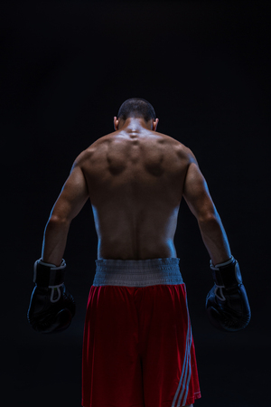 Rear view of strong young male boxer. Fitness male model wearing boxing gloves standing on black background.の写真素材