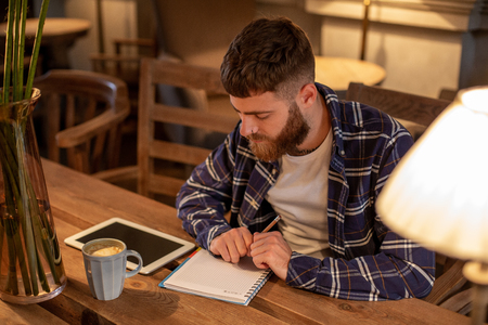 Young bearded businessman sits in cafe, home at table and writes in notebook, near lies tablet computer with black screen. Man is working, studying.の写真素材