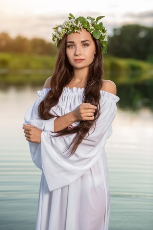 Beautiful black haired girl in white vintage dress and wreath of flowers standing in water of lake. Fairytale story. Warm art work. Sun flareの写真素材