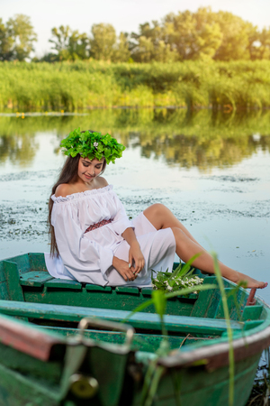 The nymph with long dark hair in a white vintage dress sitting in a boat in the middle of the river.の写真素材