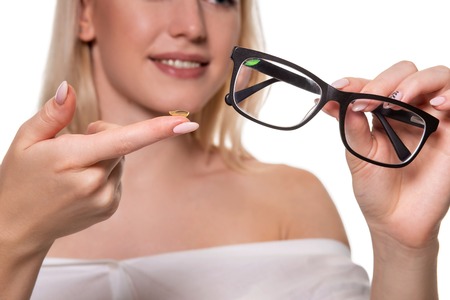 Young blonde woman holding contact lens on finger in front of her face and holding in her other hand a black glasses on white background. Close-upの写真素材
