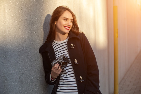 Smiling female photographer in jacket standing in front of wall ready to make new photo. Sun flare