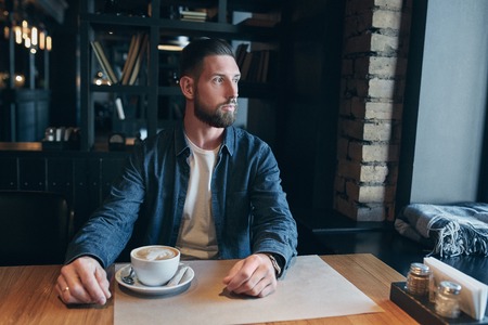 Confident man enjoying a cup of coffee while having work break lunch in indoors cafe looking pensive