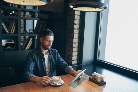 Young man drinking coffee in cafe and using tablet computerの写真素材