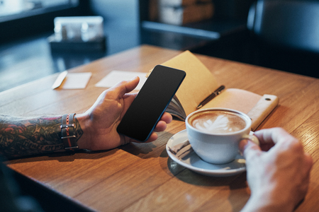 Man hands in denim shirt slide with finger on screen his smart phone, near cup with coffee on wooden tableの写真素材