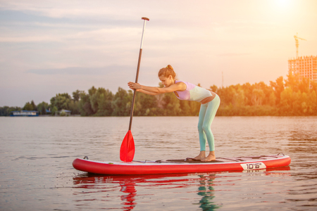 Sporty woman in yoga position on paddleboard, doing yoga on sup board, exercise for flexibility and stretching of musclesの写真素材