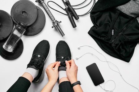Young woman in sporting leggings laces sneakers, preparing for training. Accessories for sports, bottle of water, smartphone, headphones, fitness bracelet, dumbbells on white background flat lay top view. Fitness conceptの写真素材