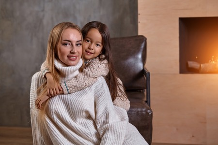 Beautiful mother woman in white sweater hugs her little daughter. Cheerful playful mood and love inside the family.の写真素材