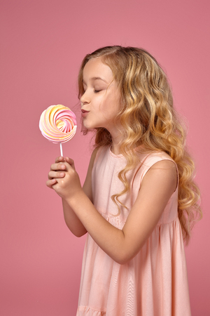 Little girl with a blond curly hair, in a pink dress is posing with a candyの写真素材