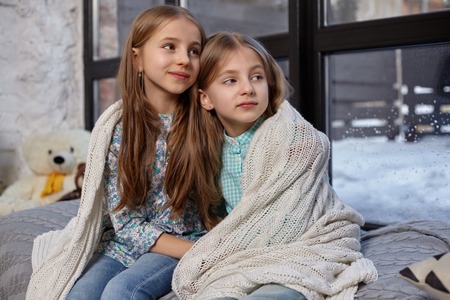 The image of two cute little sisters sitting on windowsill, covered by blanket, hugging each otherの写真素材