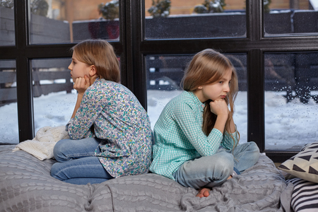 Close up photo of two cute offended little sisters who sit with their backs to each other on windowsillの写真素材