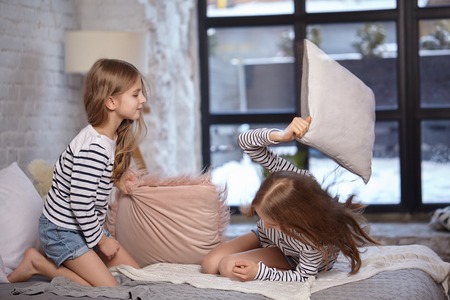 The image of two little sisters sitting on the bed in the room. They using pillow to fight each...