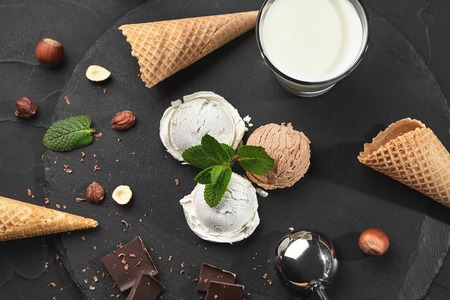 Top view of sophisticated creamy and chocolate ice cream decorated with mint, a glass of milk, metal scoop, waffle cones with chocolate and hazelnuts nearby, served on a stone slate over a black background. Close-up.の写真素材