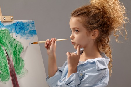 Charming school girl is painting with a watercolor brush on an easel, standing on a gray background.の写真素材