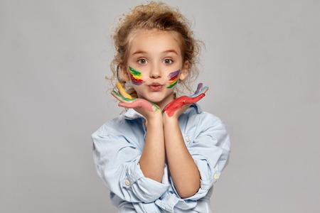 Beautiful little girl with a painted hands and cheeks is posing on a gray background.の写真素材