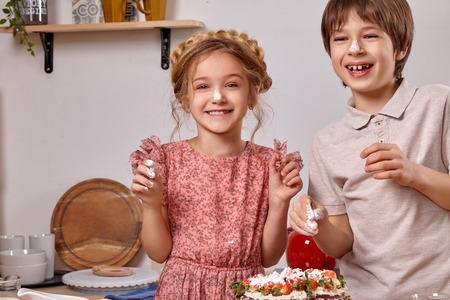 Little friends are making a cake together at a kitchen against a white wall with shelves on it.の写真素材