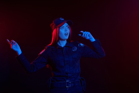 Close-up shot of a redheaded female police officer is posing for the camera against a black background with red and blue backlighting.の写真素材