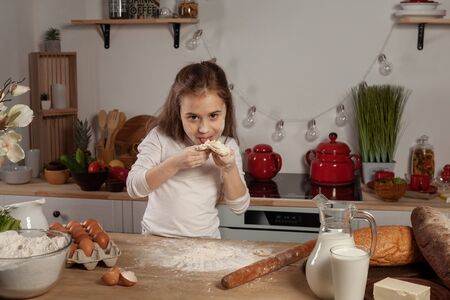 Beautiful little girl dressed in a white blouse is making a dough for baking a bread at a kitchen.