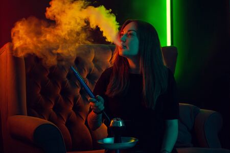 Pretty girl in a black t-shirt is smoking a hookah exhaling a smoke at a luxury night club.