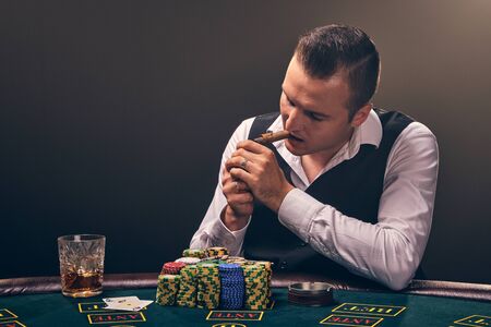 Wealthy man is smoking a cigar and playing poker with an excitement at a casino on black background.
