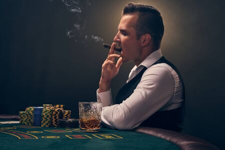 Wealthy man is smoking a cigar and playing poker with an excitement at a casino on black background.