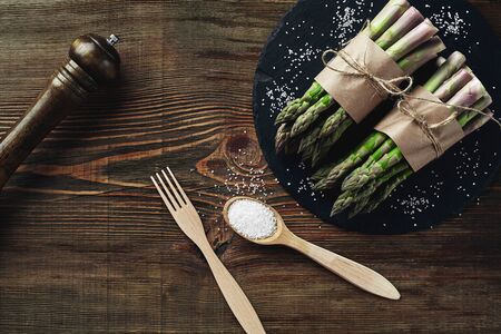 Bunches of an edible, appetizing spears of asparagus on a stone slate, wooden background. Fresh, green vegetables with seasonings, fork and spoon, kitchen pestle, top view. Healthy eating. Fall harvest, agricultural farming concept.の写真素材
