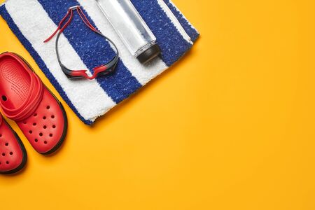 Flat lay, pool accessories such as swimming goggles, striped towel, red slippers and bottle for water on a yellow background. Multicolored beach wear. Copy space, top view, close-up.の写真素材