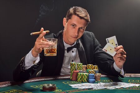 Handsome emotional man is playing poker sitting at the table in casino against a white spotlight.の写真素材