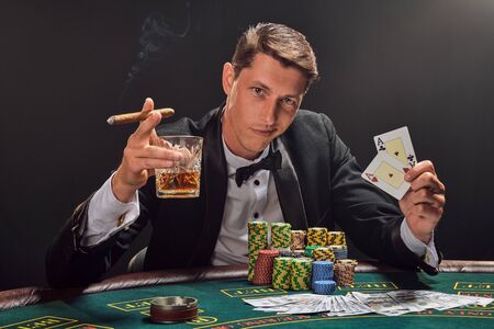 Handsome emotional man is playing poker sitting at the table in casino against a white spotlight.の素材 [FY310127508712]