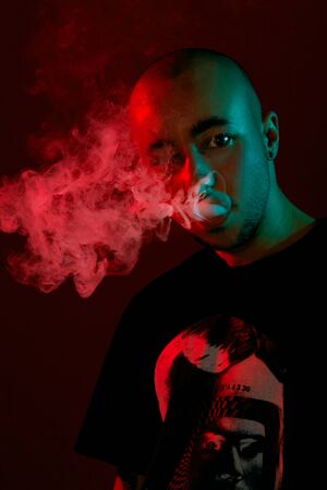 Close-up studio shot of a young bald guy vaping, blowing out a cloud of smoke on red background.
