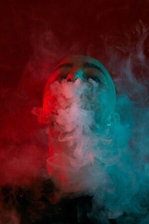 Close-up studio shot of a young bald guy vaping, blowing out a cloud of smoke on red background.の写真素材